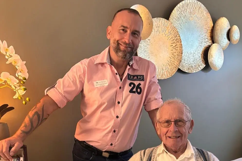Bernard Carret-Ollivier, debout, pose la main sur le dossier du fauteuil où un homme âgé est installé ; tous deux sourient.